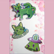A photo of three stacks of stickers, each featuring an illustration of an alien cat on a glitter background by Ayumutan Art